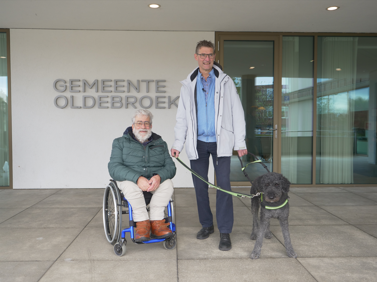 Ton van Weerdenburg met geleidehond Sofie en rolstoelgebruiker Jan Oosterbroek voor de gevel van het gemeentehuis in Oldebroek.