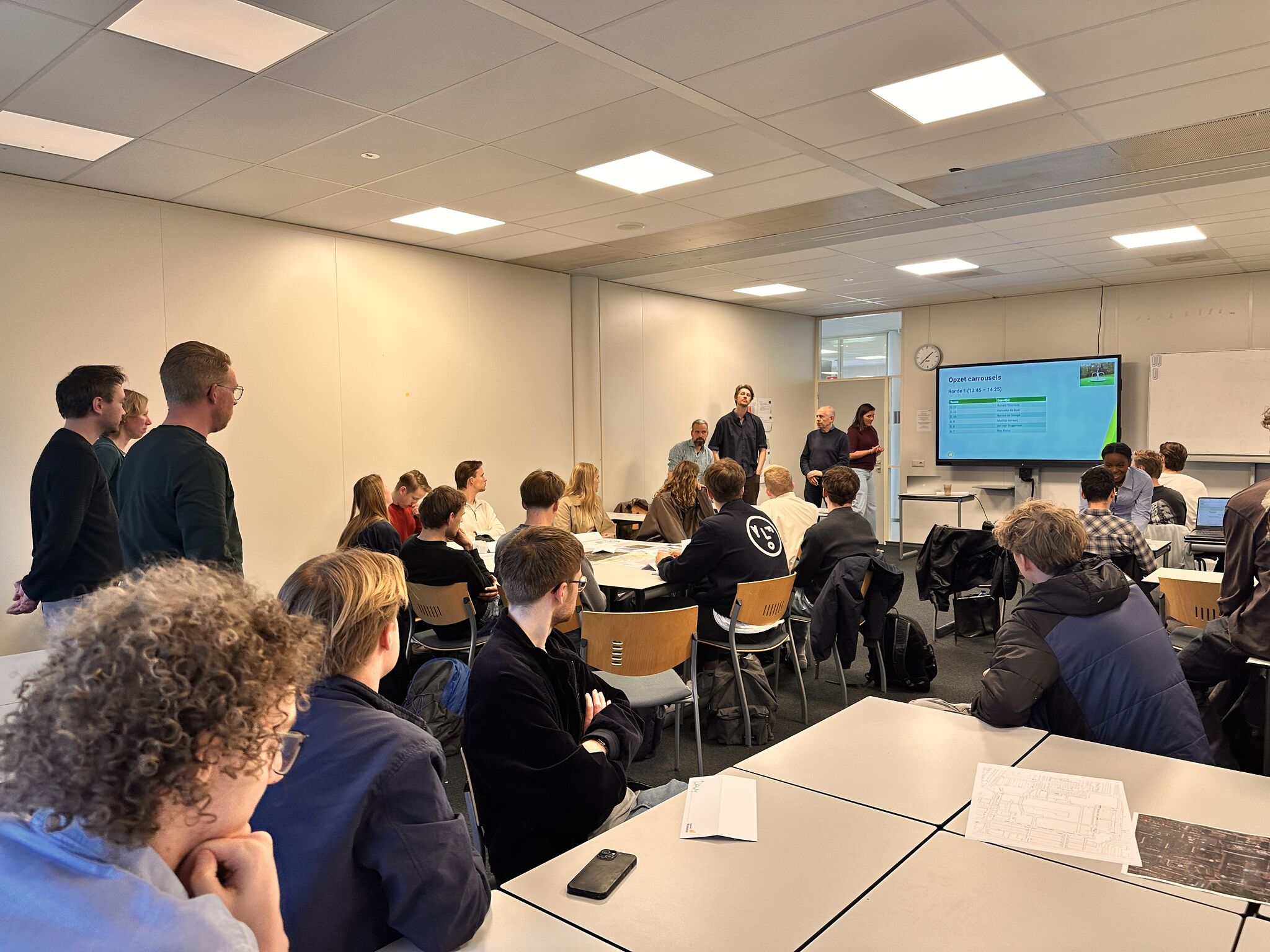 Foto van de leerlingen in het lokaal. Ze kijken naar de docent die iets aan het uitleggen is.