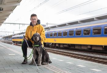 Foto van ervaringsdeskundige op het perron met hulphond en taststok