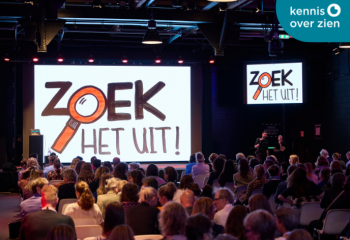 Groep mensen in zaal voor scherm met groot het Zoek het uit logo. Logo Kennis Over Zien.