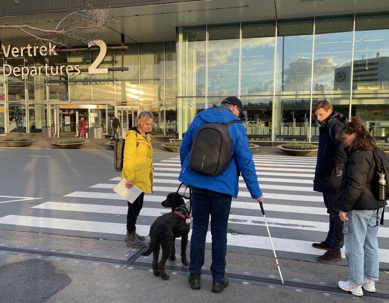 Frouck de Boer en Peter Waalboer staan voor een zebrapad bij een geleidelijn voor Vertrekhal 2 van Schiphol. Ze praten tegen twee andere mensen.