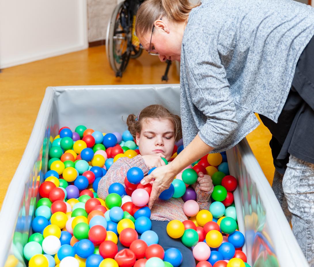 Meisje met een visuele en verstandelijke beperking in een ballenbad. Begeleidster staat ernaast en duwt ballen in de richting van de hand van het meisje.