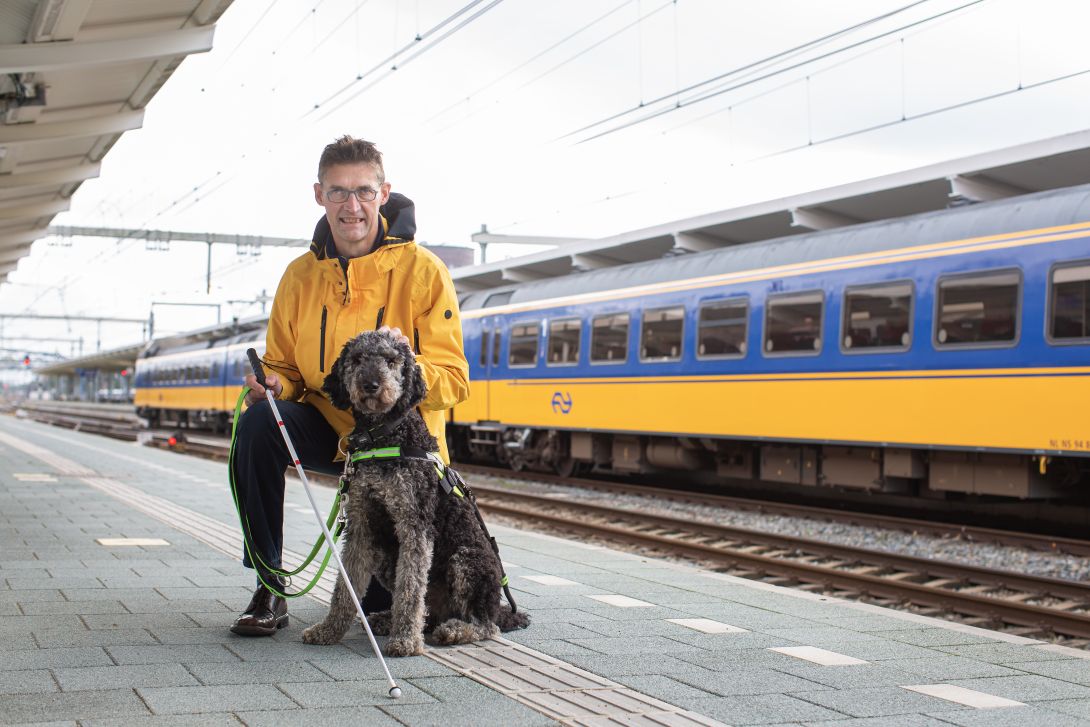 Foto van ervaringsdeskundige op het perron met hulphond en taststok